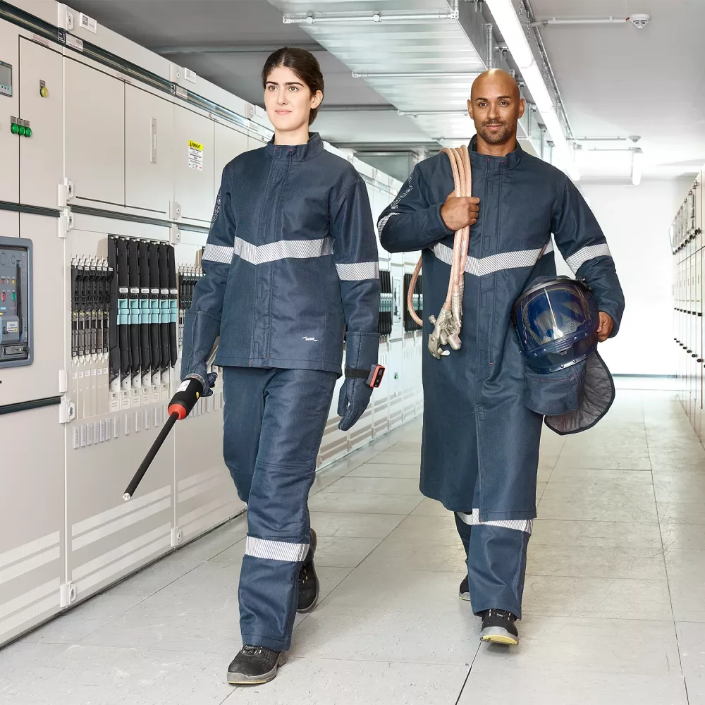 Two technicians in protective clothing walking through an electrical switchgear room, carrying insulated tools and safety equipment.