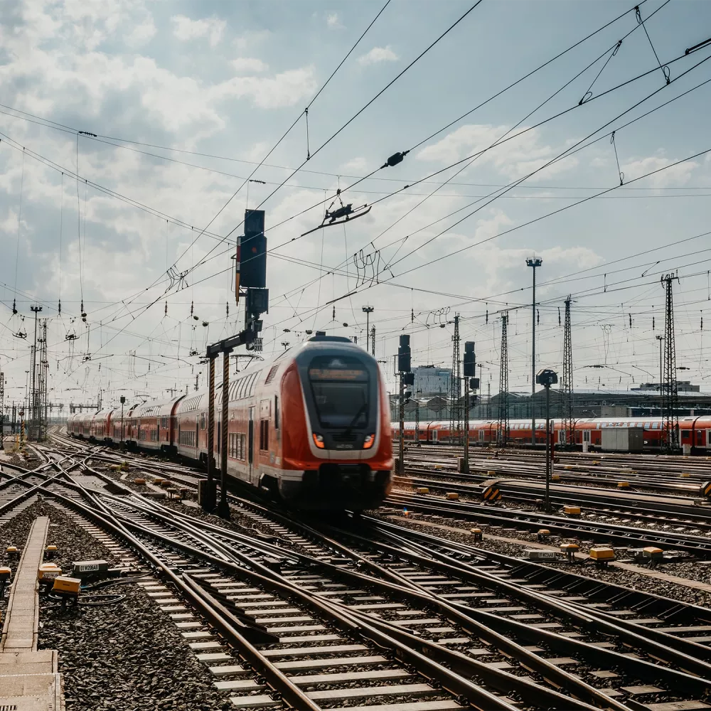 High-speed passenger train traveling through a rail yard with multiple tracks, overhead power lines, and signal systems under a cloudy sky.