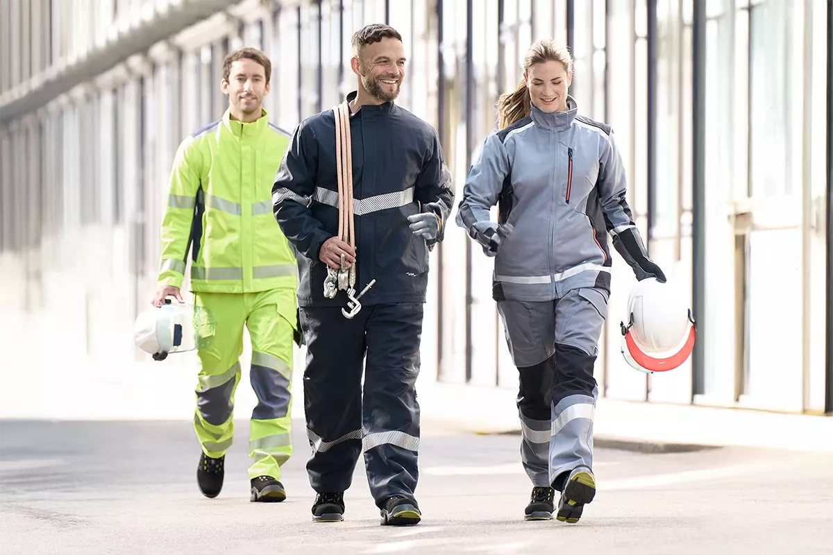 Three technicians in protective workwear walking through an industrial facility, carrying helmets and safety equipment.